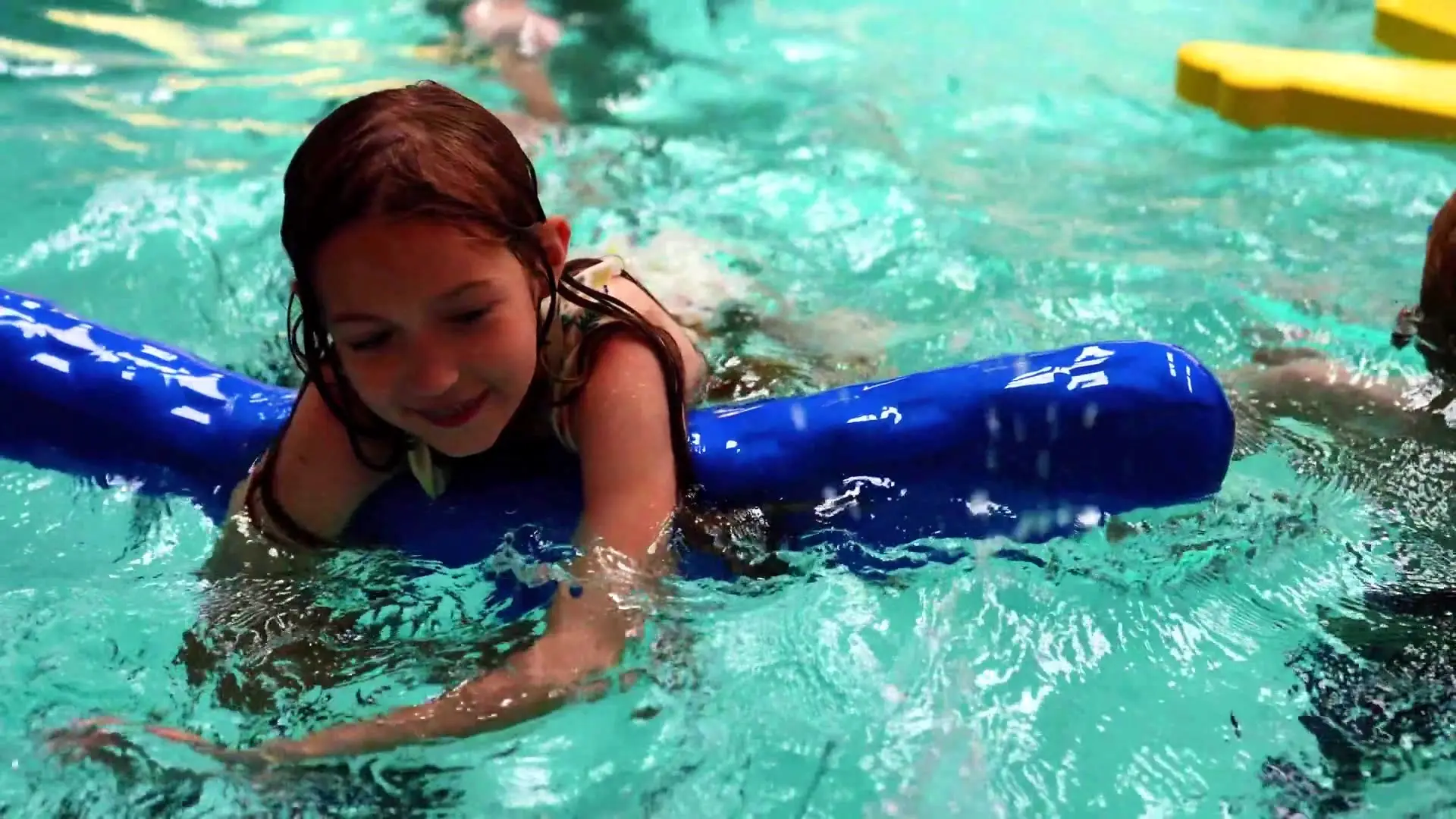 A young swimmer laughs while floating over a blue pool noodle during a party swim